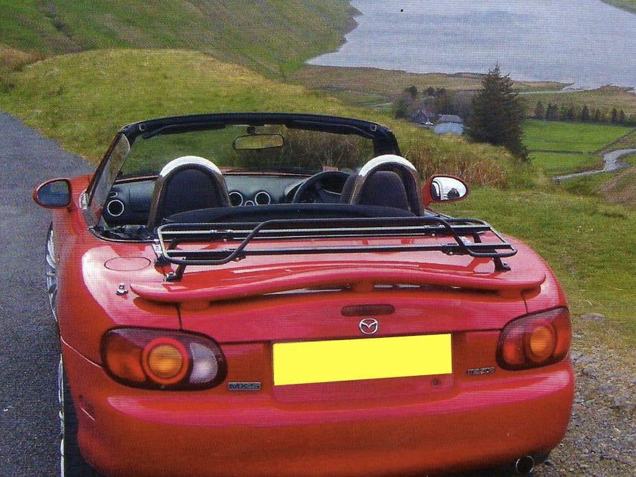 red mx5 mk2 with rear spoiler and a black boot rack fitted to the boot at the top of hill near a lake with the hood down.