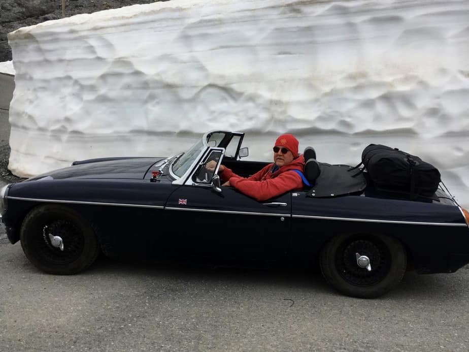 black mgb with the hood down and a drive in a red jacket and bobble hat next to a huge show wall with a boot-bag vacation boot rack fitted to the boot