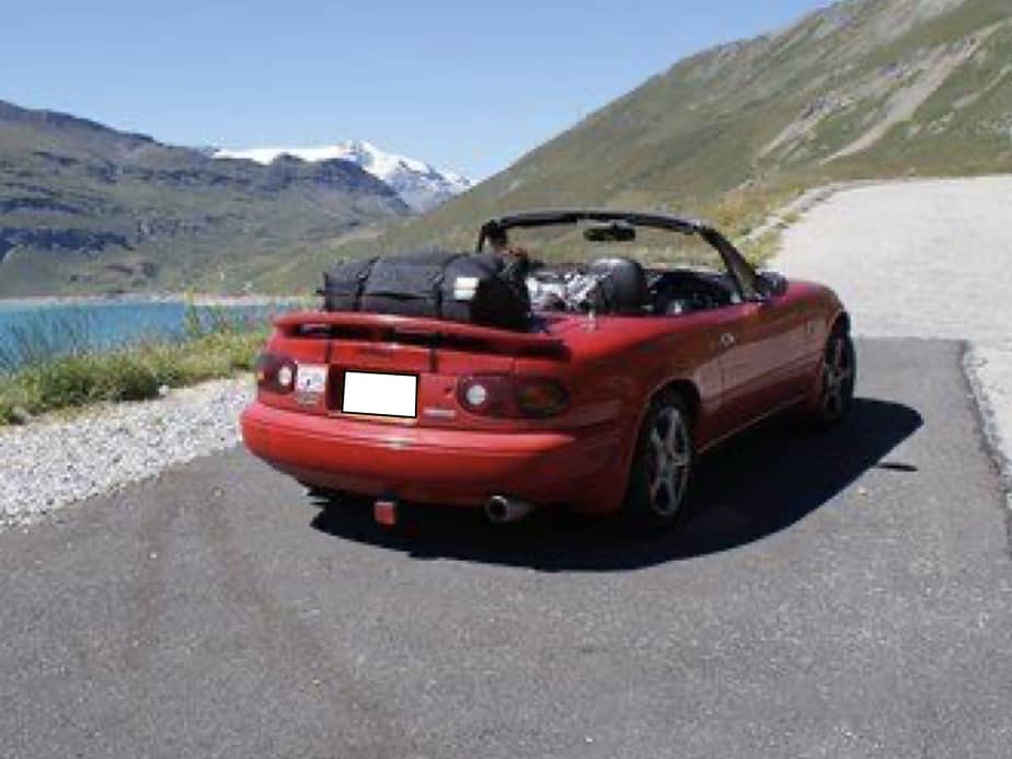 red mazda mx5 mk1 on a slope next to a lake with mountains in the background and a boot rack fitted